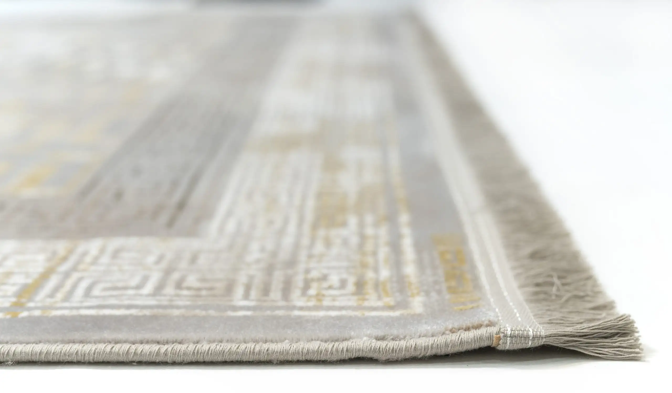 A close-up of a white and gold patterned rug with a fringe on one edge, made by Qaleen Carpets