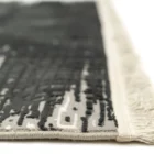 A close-up of a rug with a fringe border, made by Qaleen Carpets, featuring a pattern of dark and light grey, with a white
