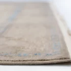 A close-up of a beige rug with a subtle blue pattern, with a fringe on the right side, made by Qaleen Carpets