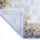 A corner of a Qaleen Carpets rug with a white background, featuring a mix of gold, blue, and brown patterns, with a fringe
