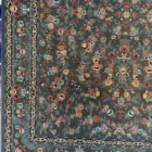 A close-up of a Qaleen Carpets rug with a floral pattern and a dark grey background, featuring a border and a fringe at the